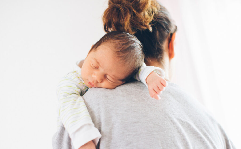 Sleeping dark haired infant sleeps over mother's shoulder while mother stand with back to camera.