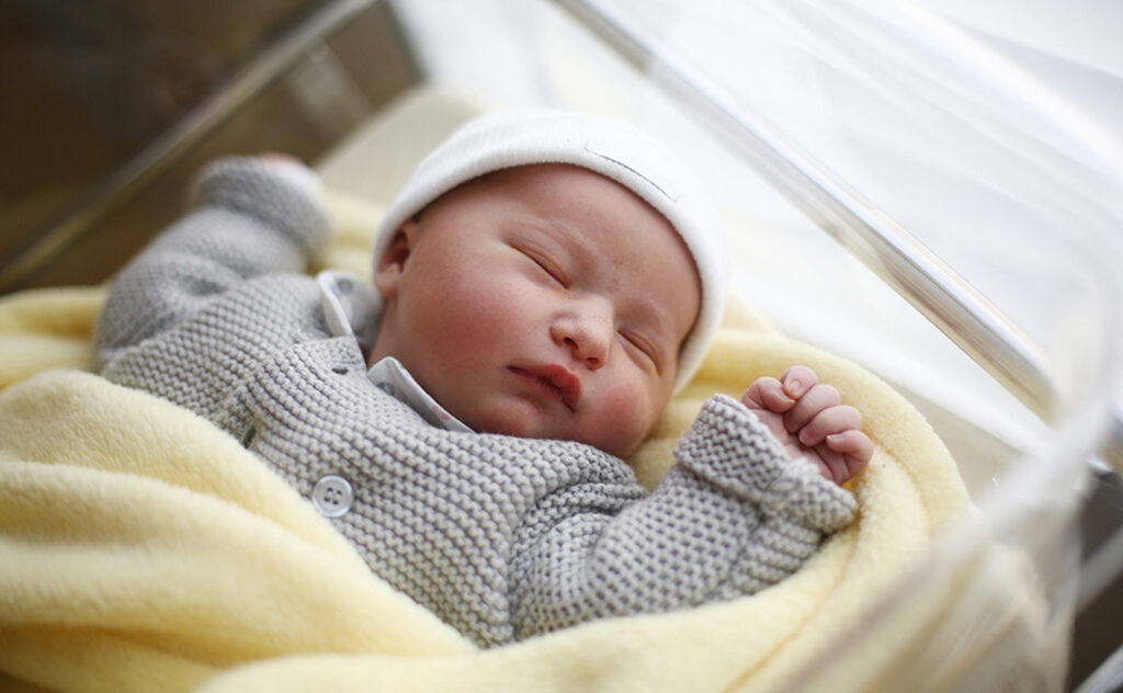 Sleeping infant in grey woollen cardigan, white beanie wrapped in lemon yellow blanket.