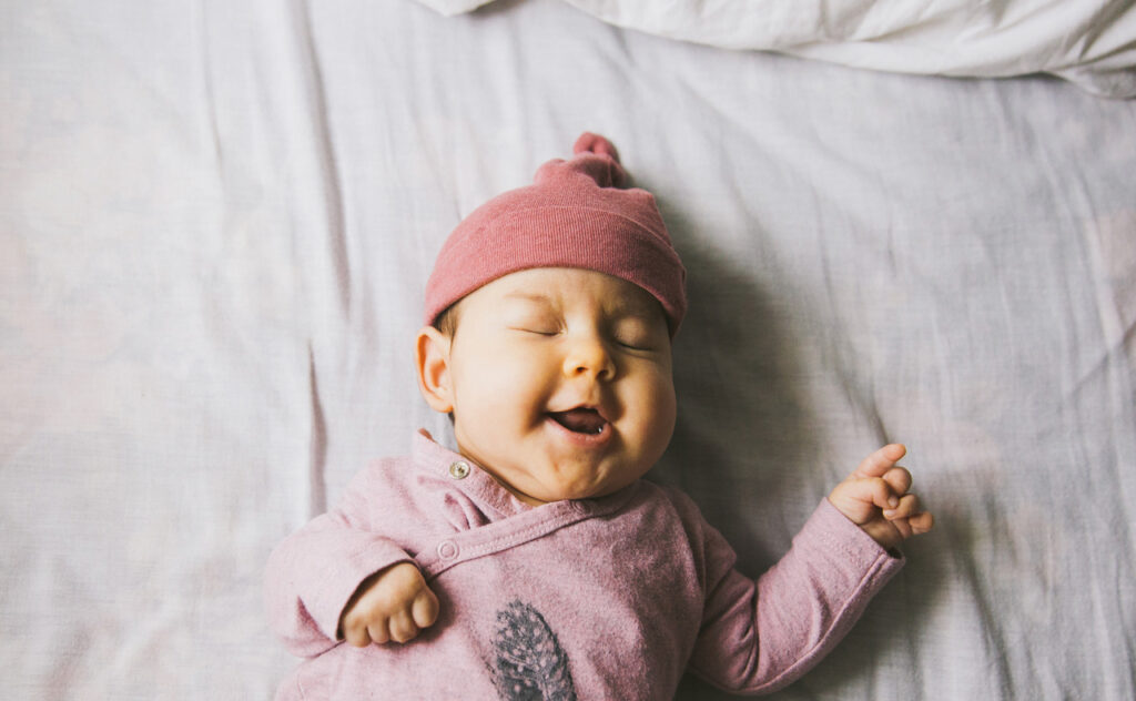 Smiling newborn with eyes closed and oink beanie wearing wrap onesie.