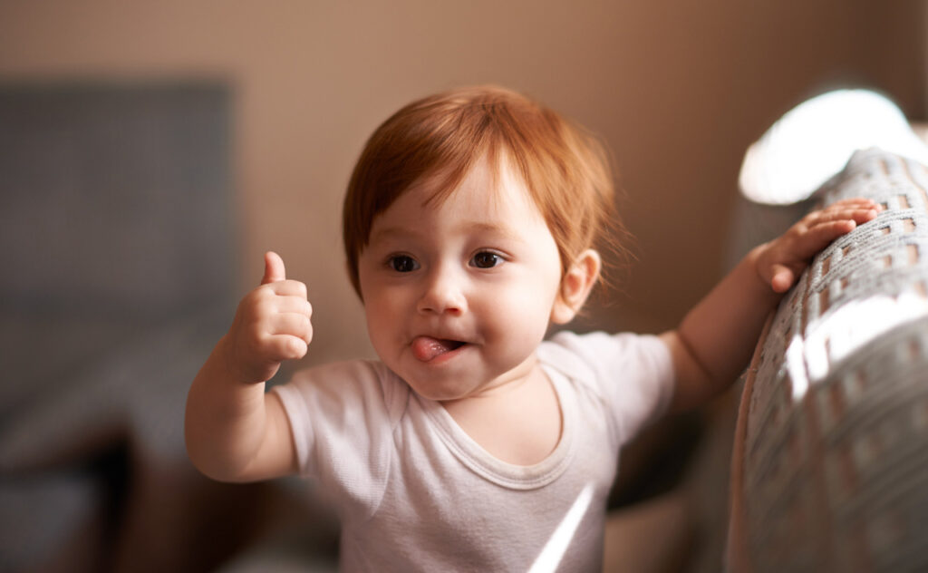 Cheeky red haired toddler with tongue out gives a thumbs up.