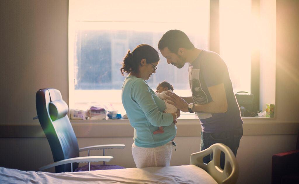 New parents stand in front of sun streamed hospital room window while staring at their newborn.