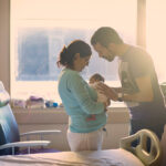 New parents stand in front of sun streamed hospital room window while staring at their newborn.