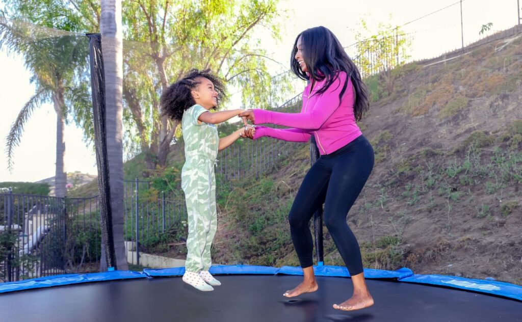 Womean with long dark hair, bright pink top and bare feet holds hands with young girl in cute camo outfit as they jump together on a trampoline