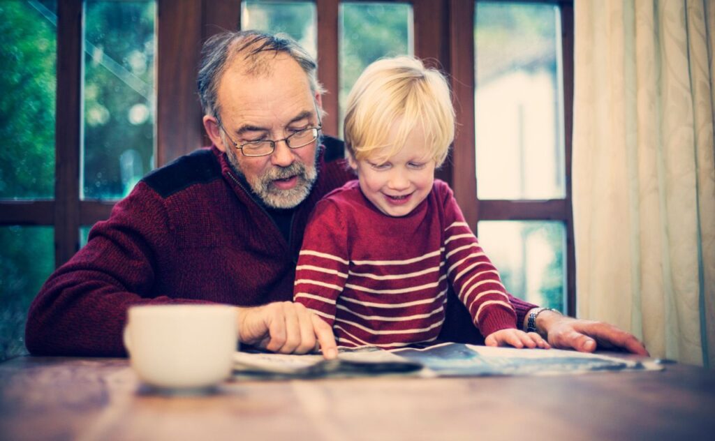 Granpda reading the newspaper to a smiling blonde boy in a red and white striped jumper