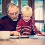 Grandad sitting with blonde haired toddler boy pointing at a book as if reading to them