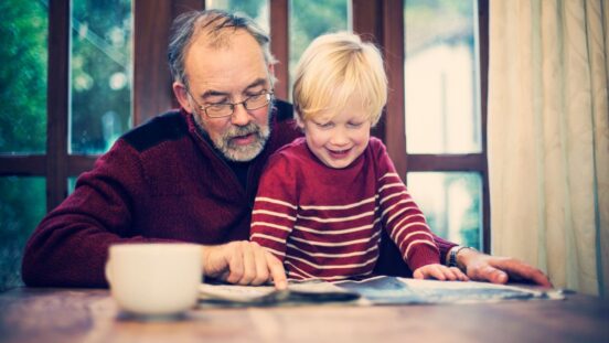 Grandad sitting with blonde haired toddler boy pointing at a book as if reading to them