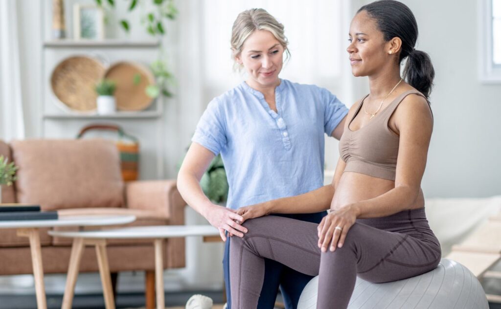Dark haired pregnant woman in exercise clothes sits on an exercise ball as a woman in a pale blue top looks at her posture