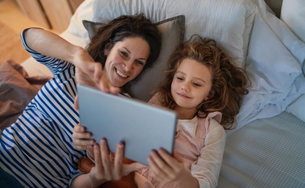 Woman with dark hair lies beside young girl and points at tablet