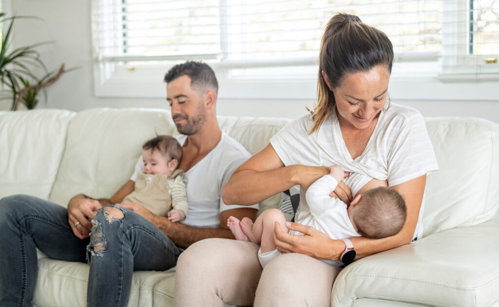 Father and mother sit on a light coloured leather lounge. The father nurses one infant while the mother breastfeeds another.