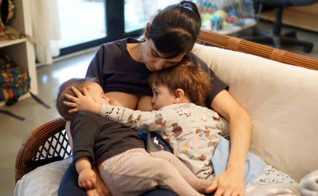 Tired mum sits on lounge tandem breastfeeding a toddler and an infant