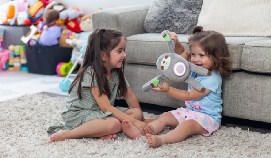 Two toddler girls sitting on the lounge room floor playing with Santi the Sloth soft toy