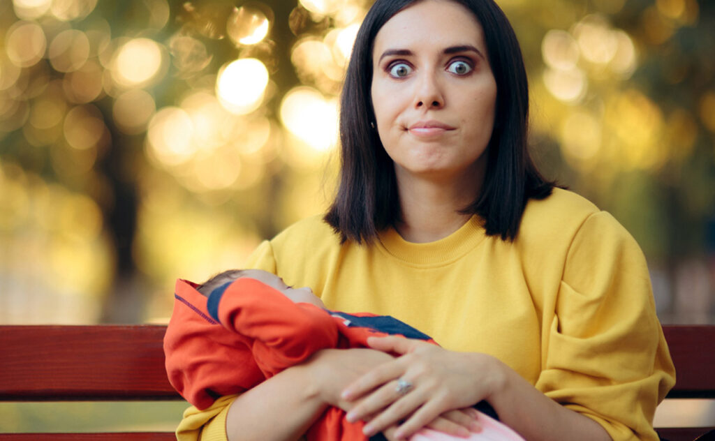 Dark haired woman wearing yellow sweater pulling a funny face while she sits on a park bench holding a baby