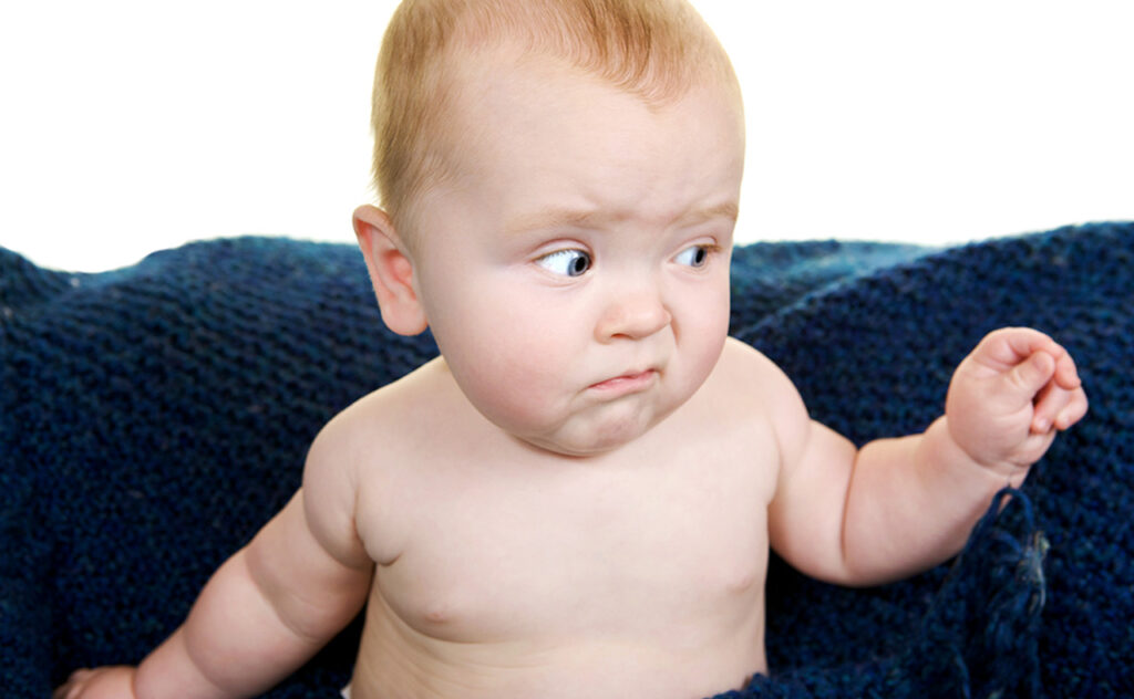 Red haired baby pulling a funny face while sitting up against a navy blue wooden blanket.