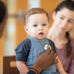 Cute toddler boy with his mouth open, looking at the camera as a doctor's hand put a stethoscope to his chest