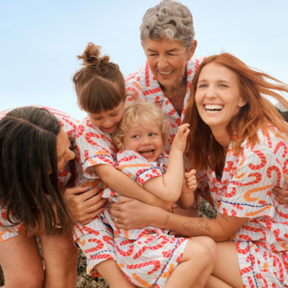 Family of women including grandmother, mother, sister and two kids cuddling and laughing wearing matching short PJs.