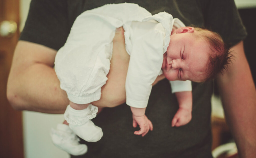 Sleeping newborn in white onesie and booties sleeping comfortably while laying over one raised adult hand.