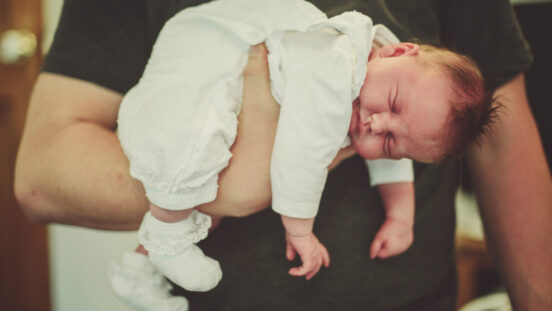 Sleeping newborn in white onesie and booties sleeping comfortably while laying over one raised adult hand.