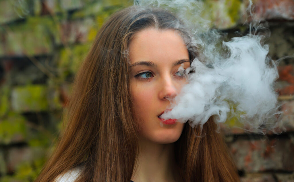 Vape teenager. Young pretty white girl in white cardigan smoking an electronic cigarette opposite destroyed brick wall on the street in the spring. 