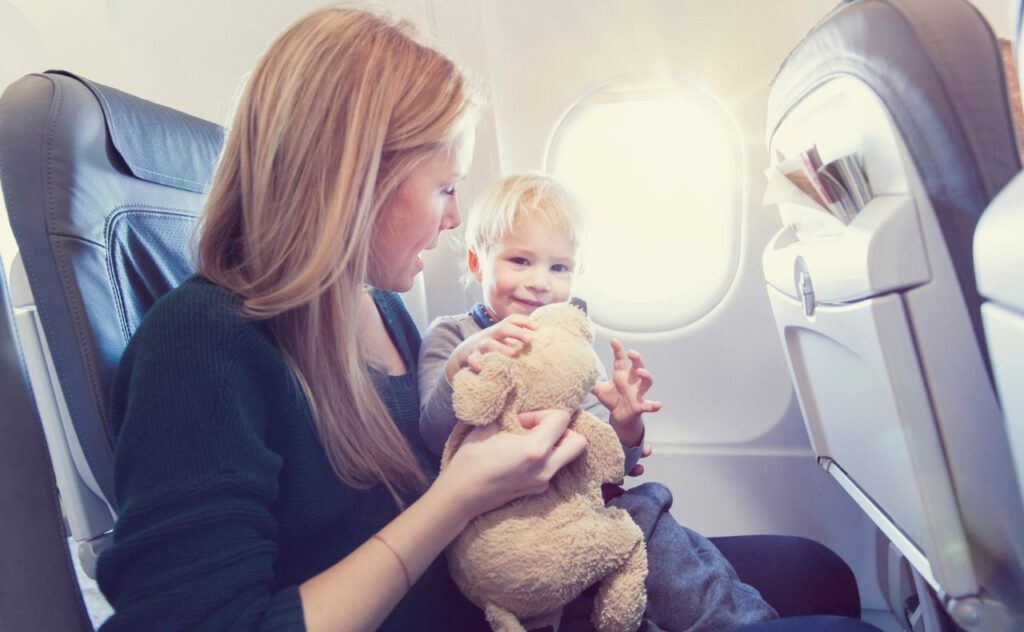 Woman with long blonde hair holding a soft brown cuddly toy for a happy blonde toddler boy sitting on her lap on a plane