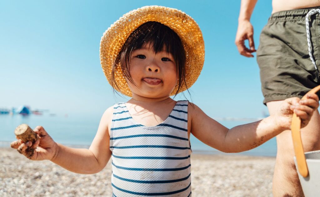 LIttle girl with dark hair and a straw hat, wearing blue and white striped swimmers at the beach