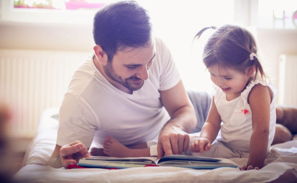 Dark haired dad in white t-short lying on the bed with a dark haired toddler with pigtails as they point at a story book