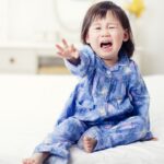 Dark haired toddler in blue pyjamas sitting on a bed, crying and reaching out with one hand