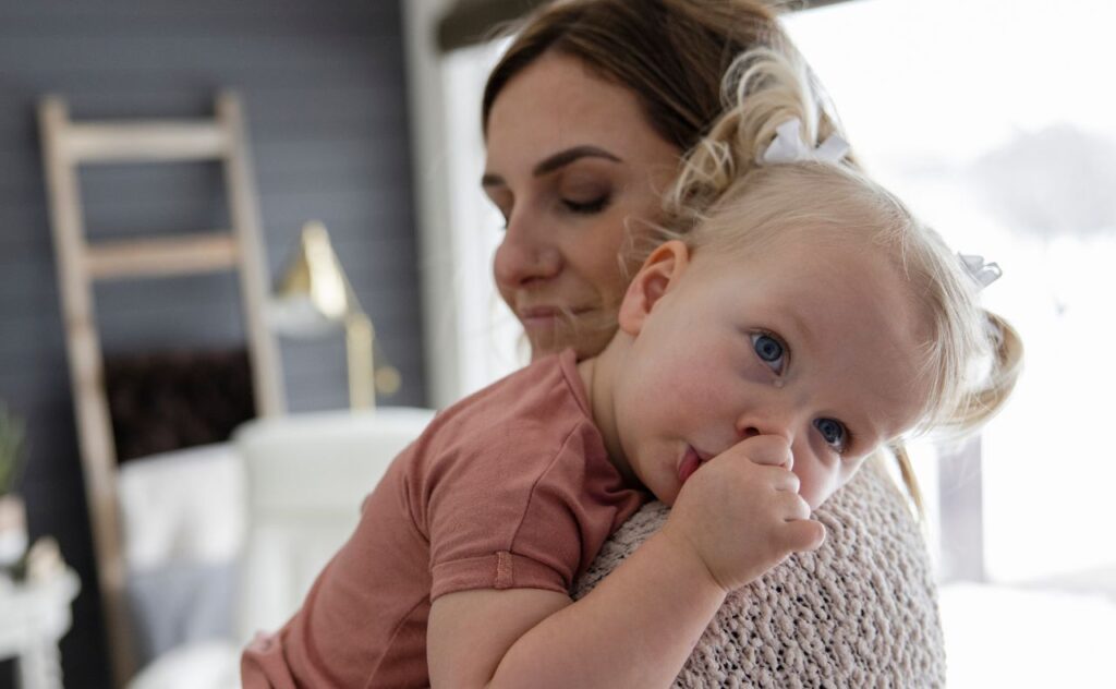 Dark haired woman cuddling toddler with blonde ponytails who is sucking her thumb