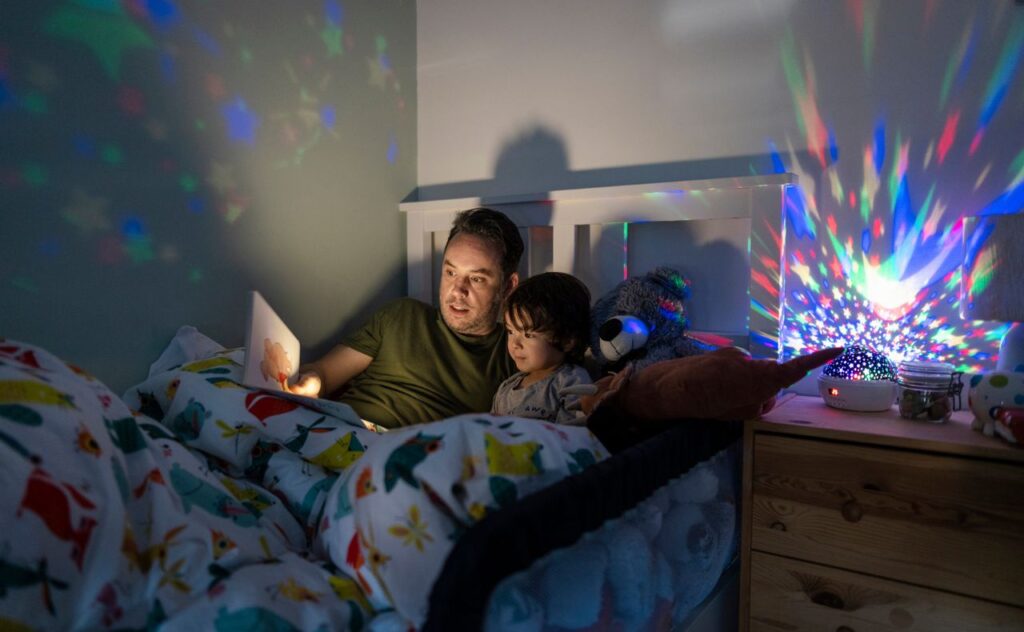 Dad sitting in a child's bed with a dark-haired toddler reading story books at bedtime