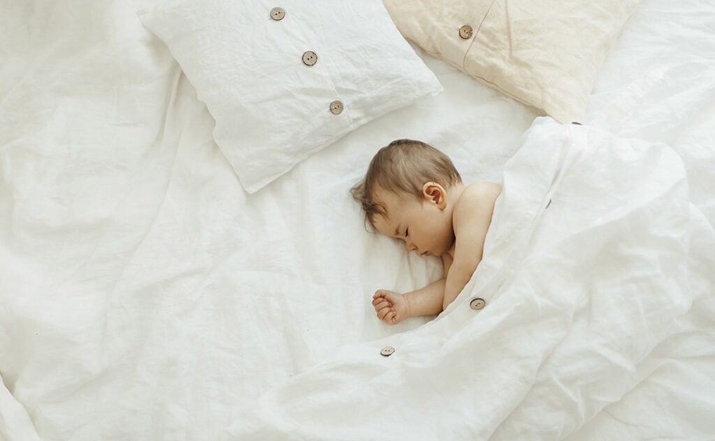 Peaceful baby lying on a white natural linen bed while sleeping in a bright room.