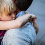 Blonde toddler girl looks upset and has arms around her dad's neck