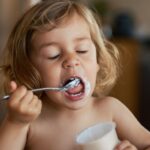 Blonde hair toddler girl eating yoghurt with her eyes closed