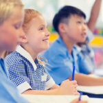 Group of children listening to the teacher. They are in a classroom. Multiethnic group with Caucasian and Asian kids. School girl is smiling and happy.