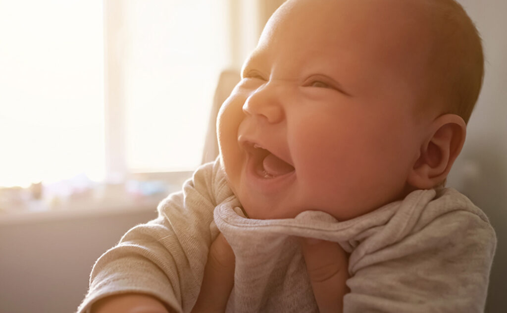Cute newborn girl with plump cheeks smiles widely feeling happily at home. Mother holds baby with hands against bright window in bedroom close view.
