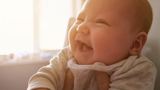 Cute newborn girl with plump cheeks smiles widely feeling happily at home. Mother holds baby with hands against bright window in bedroom close view.