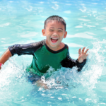 Happy Asian boy wearing a blue and green rashie splashes in the water