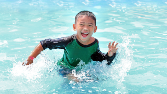 Happy Asian boy wearing a blue and green rashie splashes in the water