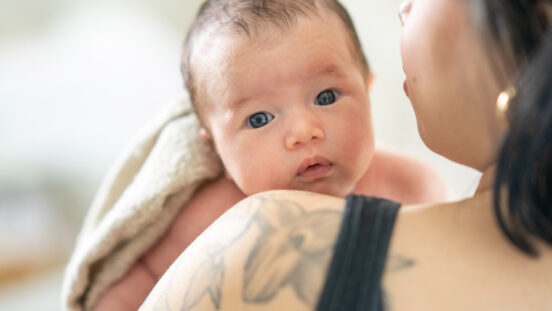 A new mother holds her infant up over her shoulder as she walks around the nursery and burps her child after feeding. The baby is skin-to-skin with her mother and lighting wrapped wth a blanket overtop.