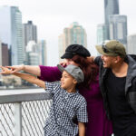 A mixed race family looking really happy pointing at something on the other side of the Brisbane river. They are on the river's egde with cityscape behind them