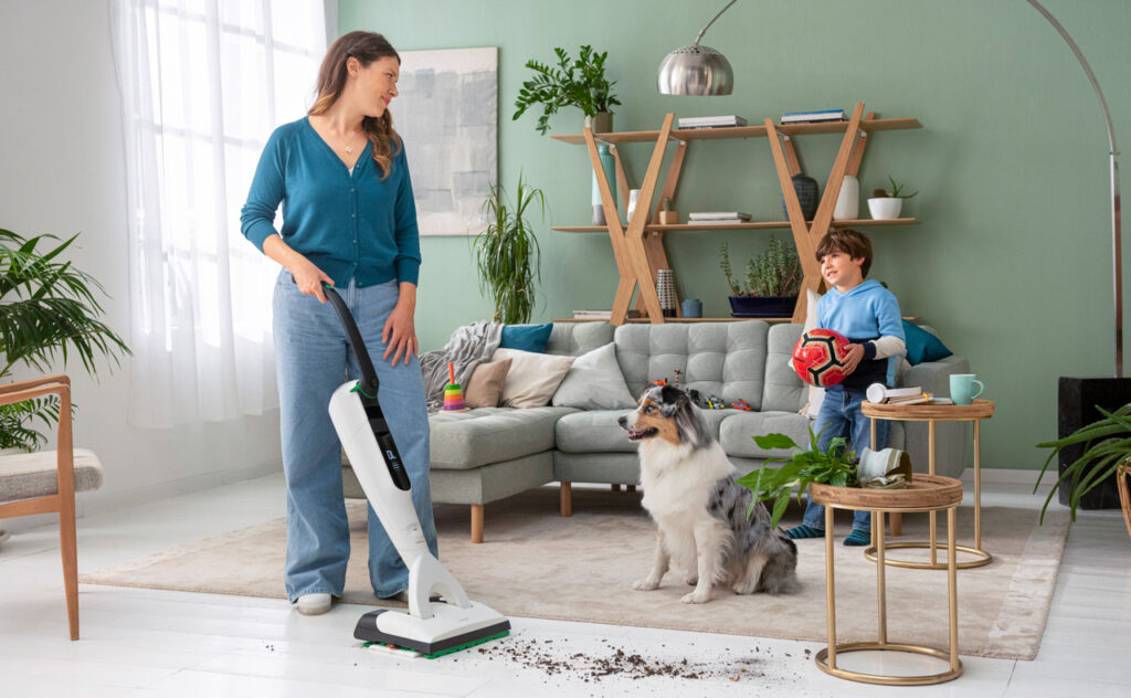 Woman uses Thermomix Kobold cordless mop vac in modern living area.= with small boy child and dog looking on.