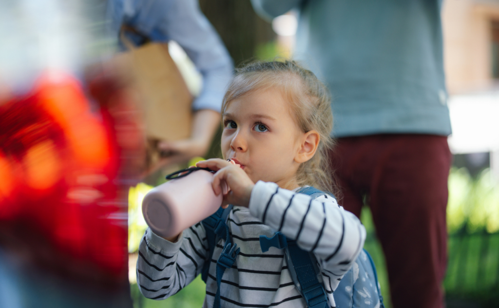 Girl with long blonde plait carrying a blue backpack and drinking from a pink water bottle
