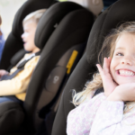 Preschooler girl smiles from her car seat as Mum buckles in smaller boy in his car seat