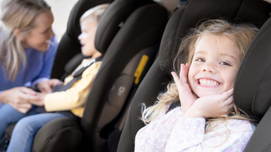 Preschooler girl smiles from her car seat as Mum buckles in smaller boy in his car seat