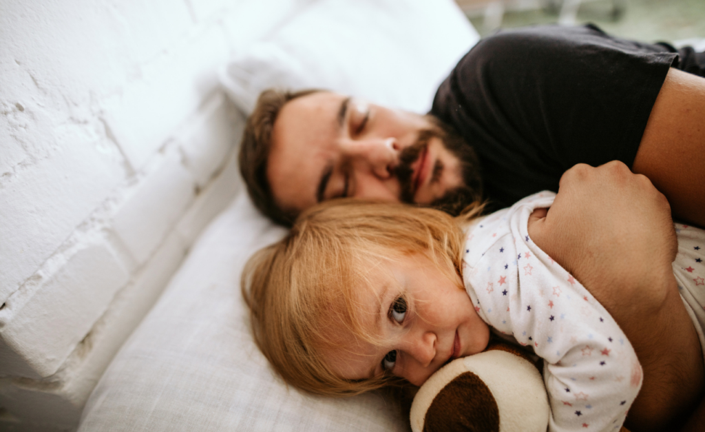 Sleeping dad cuddles blonde toddler who looks cheekily wide awake