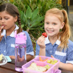 Two girls sitting at table outdoors in school uniform eating packed lunch. Girl with red hair eating strawberry and smiling towards camera