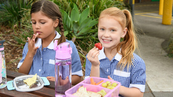 Two girls sitting at table outdoors in school uniform eating packed lunch. Girl with red hair eating strawberry and smiling towards camera