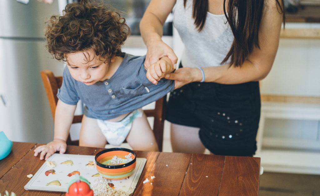 Curled haired toddler standing on chair at dining table with mum behind