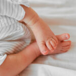 Cute sleeping newborn baby feet on white blanket. Baby in a white gender neutral romper on the bed. Little toes, heels.