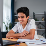 Dark hair young teen smiling at an open laptop, workbooks open on desk