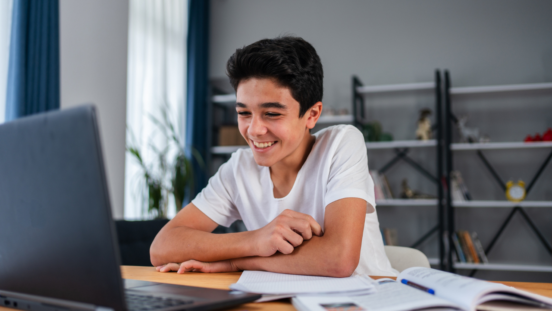 Dark hair young teen smiling at an open laptop, workbooks open on desk