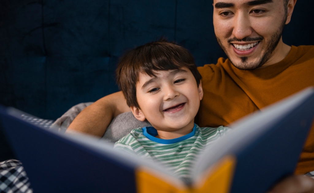 A dark haired toddler boy sits with a dark haired man, both are smilng at a book they are reading